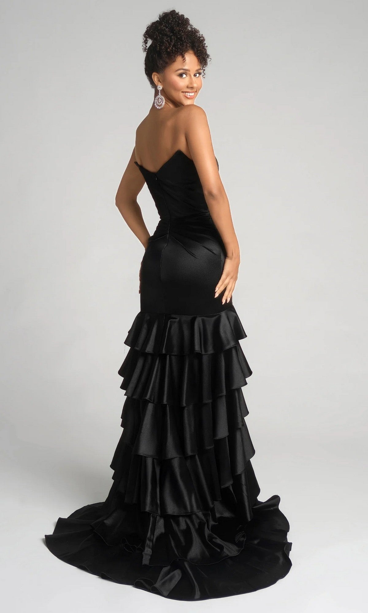 Smiling over her shoulder, a woman wears the Portia and Scarlett XO021 long prom dress—a stunning black strapless mermaid gown with a tiered ruffle skirt—posing against a plain background.